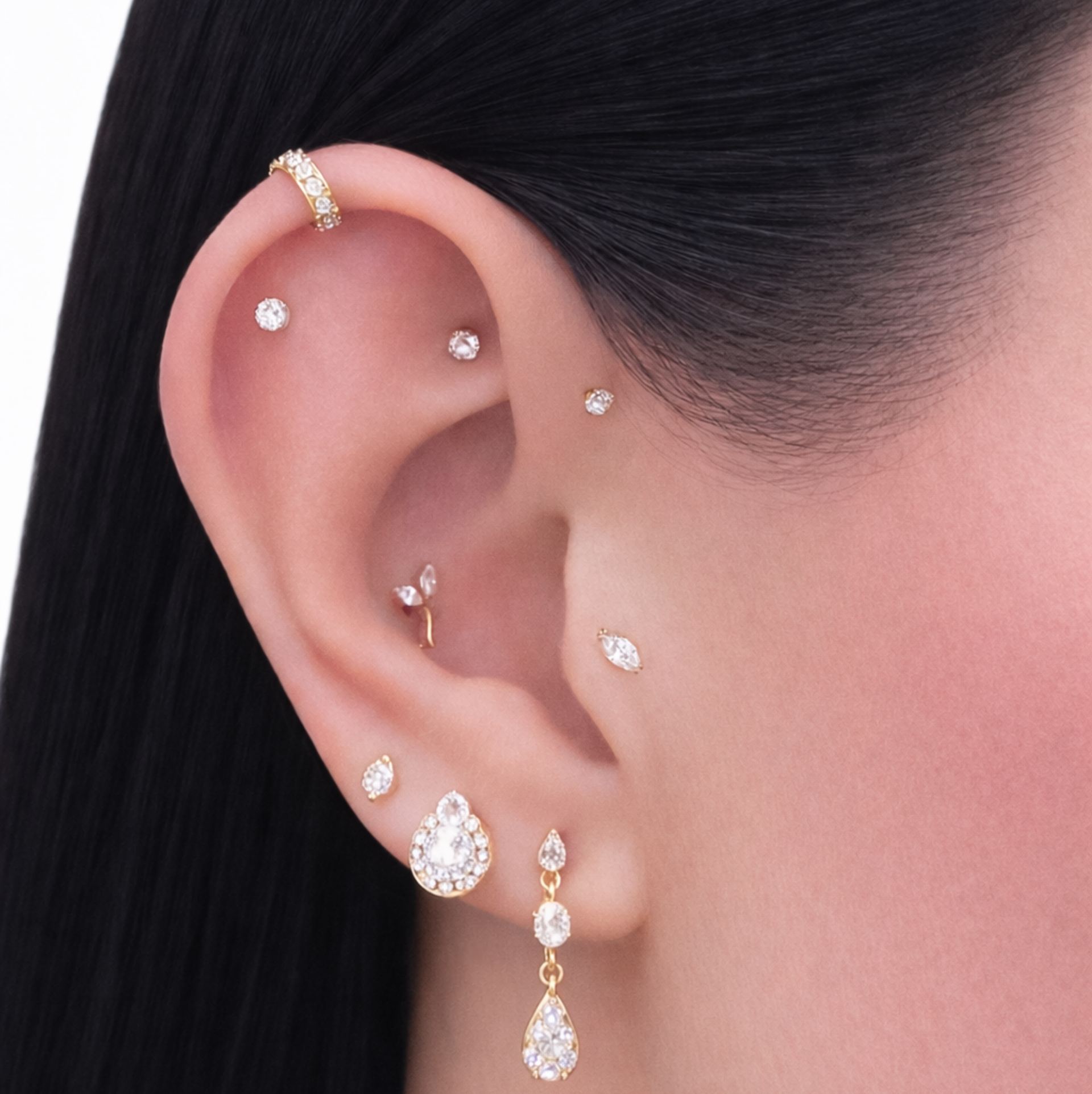 Close-up of a curated ear stack: a pave gold cuff along the outer helix, three small stud piercings along the upper ear, a flower-shaped stud and a marquise stud near the tragus and conch, and on the lobe a stud paired with a pave teardrop stud and a matching dangling teardrop drop earring