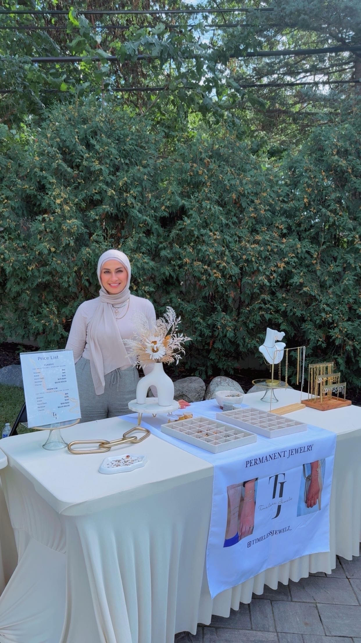Huda standing behind a Timeless Jewels pop-up market table outdoors, greenery in the background, jewelry displayed on stands, a handwritten Price List sign, a flower arrangement, and a fabric banner reading PERMANENT JEWELRY with the Instagram handle @timelessjewelz
